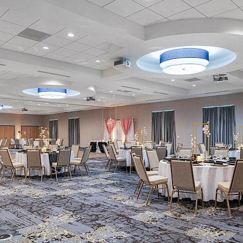 A large, elegantly decorated banquet hall with round tables set for an event, featuring modern lighting fixtures and draped wall sections.