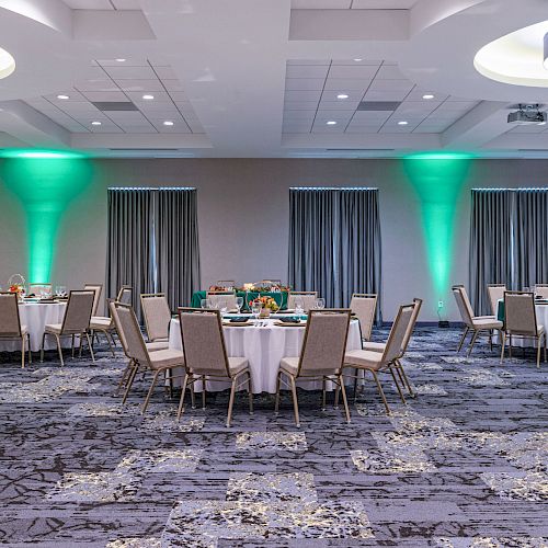 A well-lit banquet hall with round tables and beige chairs, green accent lights, and a carpeted floor.