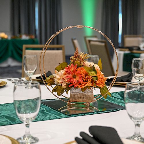 A table is elegantly set with floral centerpiece, green table runner, and glassware. Chairs and another decorated table are in the background.