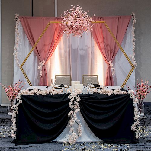 A beautifully decorated event table features a black and white cloth, pink flowers, a hexagonal arch with pink drapes, and floral arrangements around.
