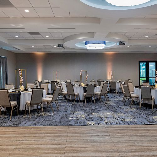 A modern banquet hall with round tables and upholstered chairs, featuring elegant lighting and decor. The room is ready for an event.