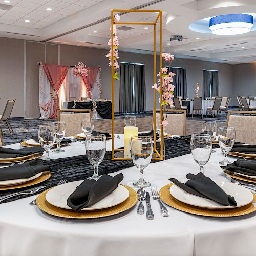 A decorated banquet room with round tables set for an event, featuring gold accents, black napkins, glassware, a candle, and a vertical floral centerpiece.