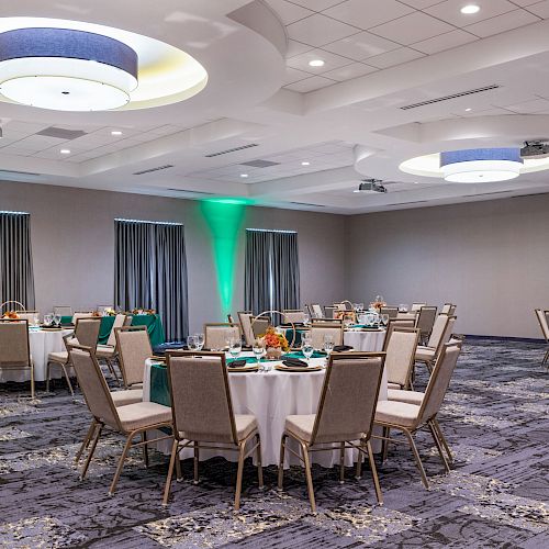 The image shows a banquet hall set up with round tables and chairs, green accent lighting, and elegant ceiling fixtures.