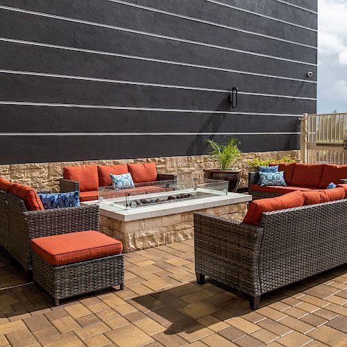 An outdoor seating area with wicker furniture, red cushions, a fire pit table, and potted plants on a paved patio in front of a black-striped wall.