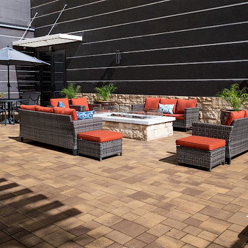 An outdoor seating area with wicker furniture and red cushions surrounds a fire pit, next to a shaded dining table and plants, on a paved patio.