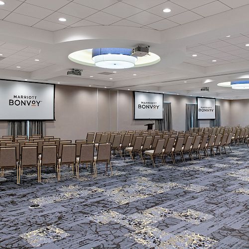 A large conference room with rows of beige chairs facing three projection screens with 