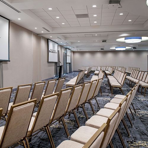 An empty conference room with rows of beige chairs facing two screens displaying the 
