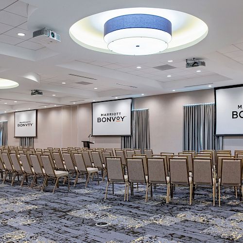 The image shows a conference room setup with rows of beige chairs facing two large screens displaying the 