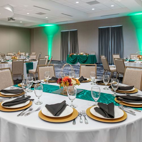 An event hall set up with round tables, each adorned with elegant tableware, black napkins, and floral centerpieces, complemented by green uplighting.
