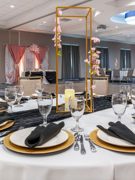 A beautifully set table with gold plates, black napkins, and glassware in an elegant banquet hall decorated with flowers and modern lighting.