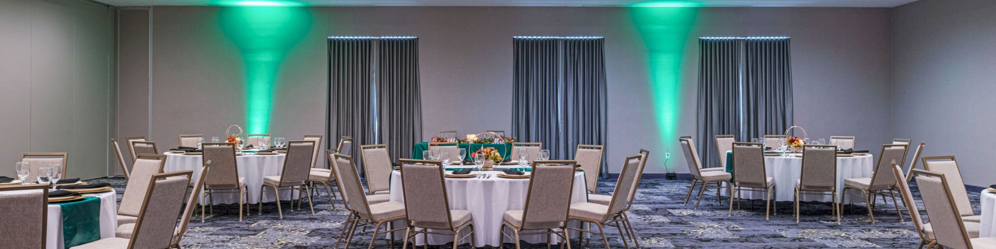 This image shows a decorated event room with round tables and chairs, subtle lighting, and green uplights on the walls.