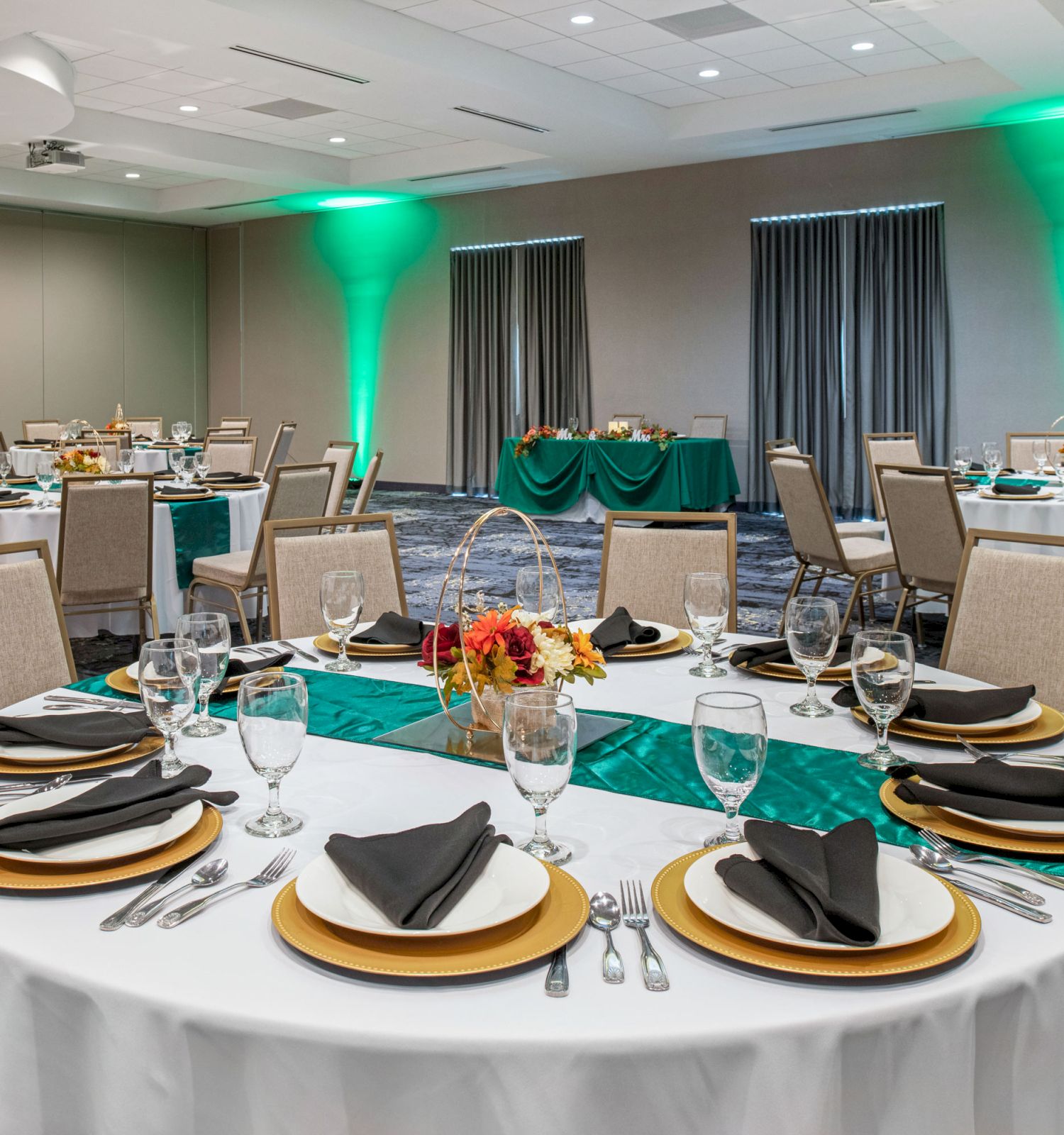 A banquet hall with round tables set with plates, glasses, cutlery, and black napkins on gold chargers, with green accents and centerpiece flowers.