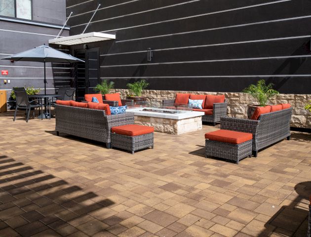 The image shows an outdoor patio area with wicker furniture, red cushions, a fire pit, potted plants, and a table with an umbrella.