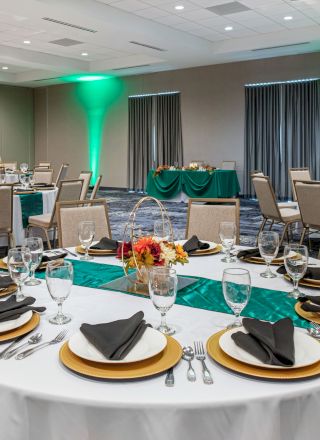 The image shows a banquet hall with round tables set for dining, adorned with centerpieces and neatly arranged tableware, with green uplighting on the walls.