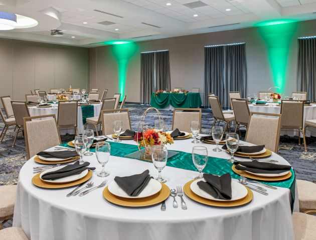 The image shows a banquet hall with round tables set for dining, adorned with centerpieces and neatly arranged tableware, with green uplighting on the walls.
