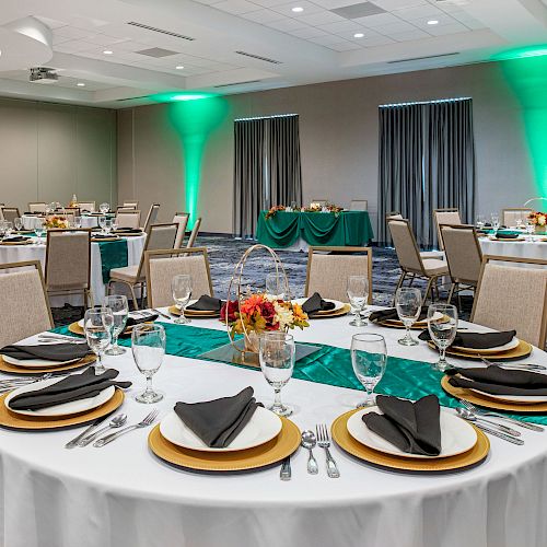 A decorated banquet hall with round tables draped in white tablecloths, set with black napkins, plates, cutlery, and glasses, and green accents.