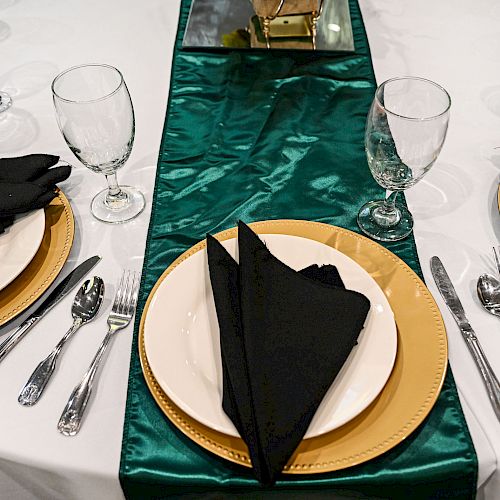 A table set for a formal event with gold-rimmed plates, black napkins, silver cutlery, and green table runner, and clear glasses on a white tablecloth.