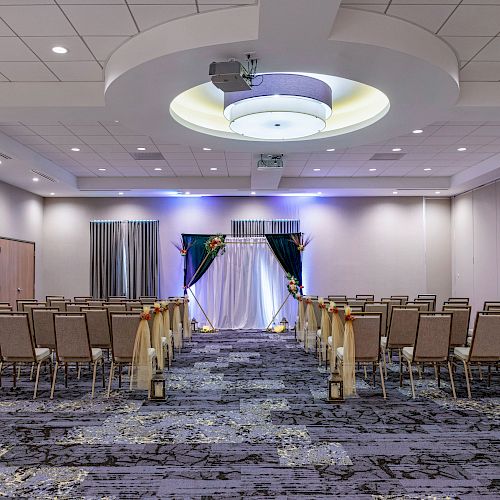 The image shows a decorated venue with rows of chairs arranged for an event, featuring a draped backdrop and spotlights in a large, carpeted room.