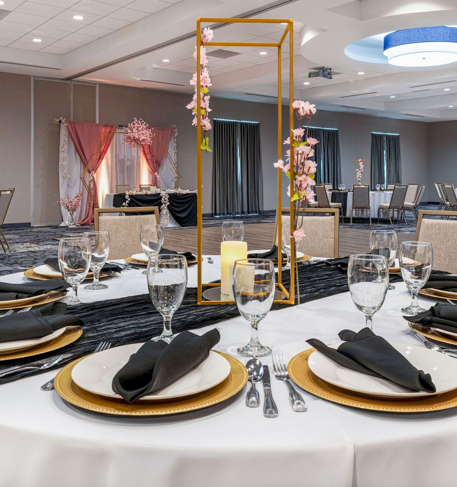 A round banquet table is set with plates, napkins, and glasses in an elegantly decorated room with other similarly arranged tables and modern lighting.